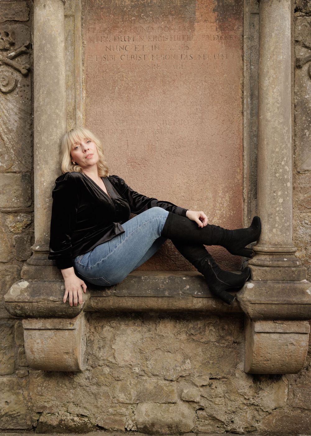 Blonde woman in black velvet top and jeans posing on stone ledge.