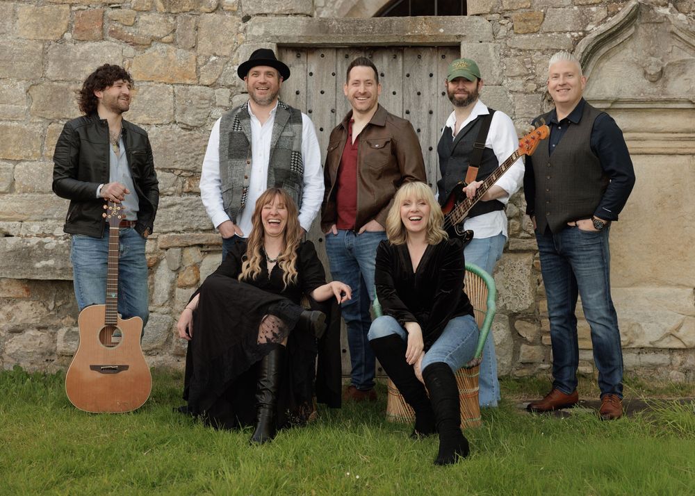 Eight-member folk band posing outdoors near stone building, holding guitars and instruments, dressed in casual attire.