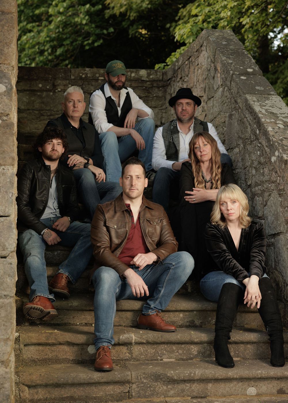 Seven-member music band poses on stone steps outdoors surrounded by lush green trees.