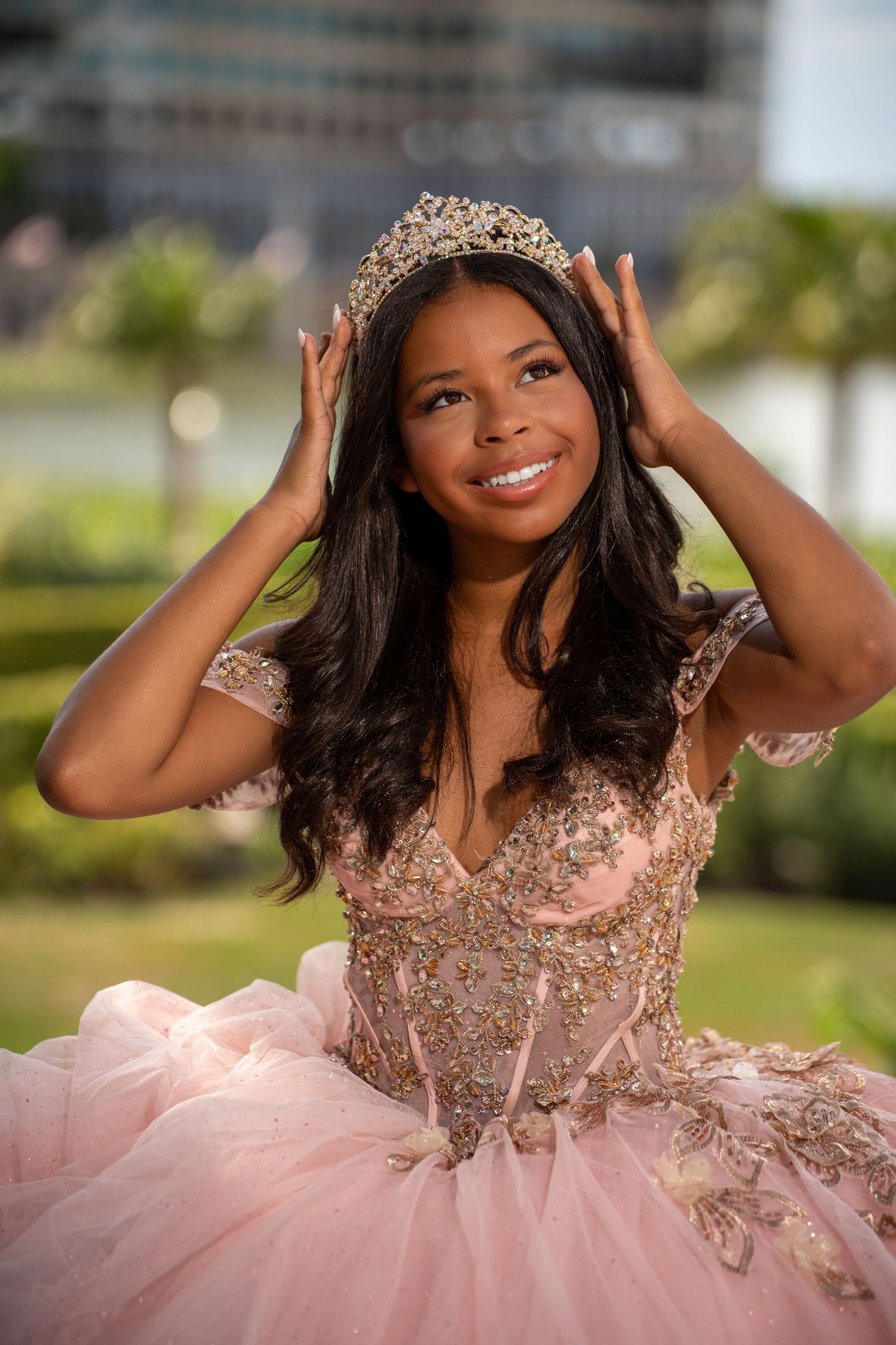 Young woman in pink quinceañera ball gown adjusts gold tiara crown while smiling outdoors at celebration.