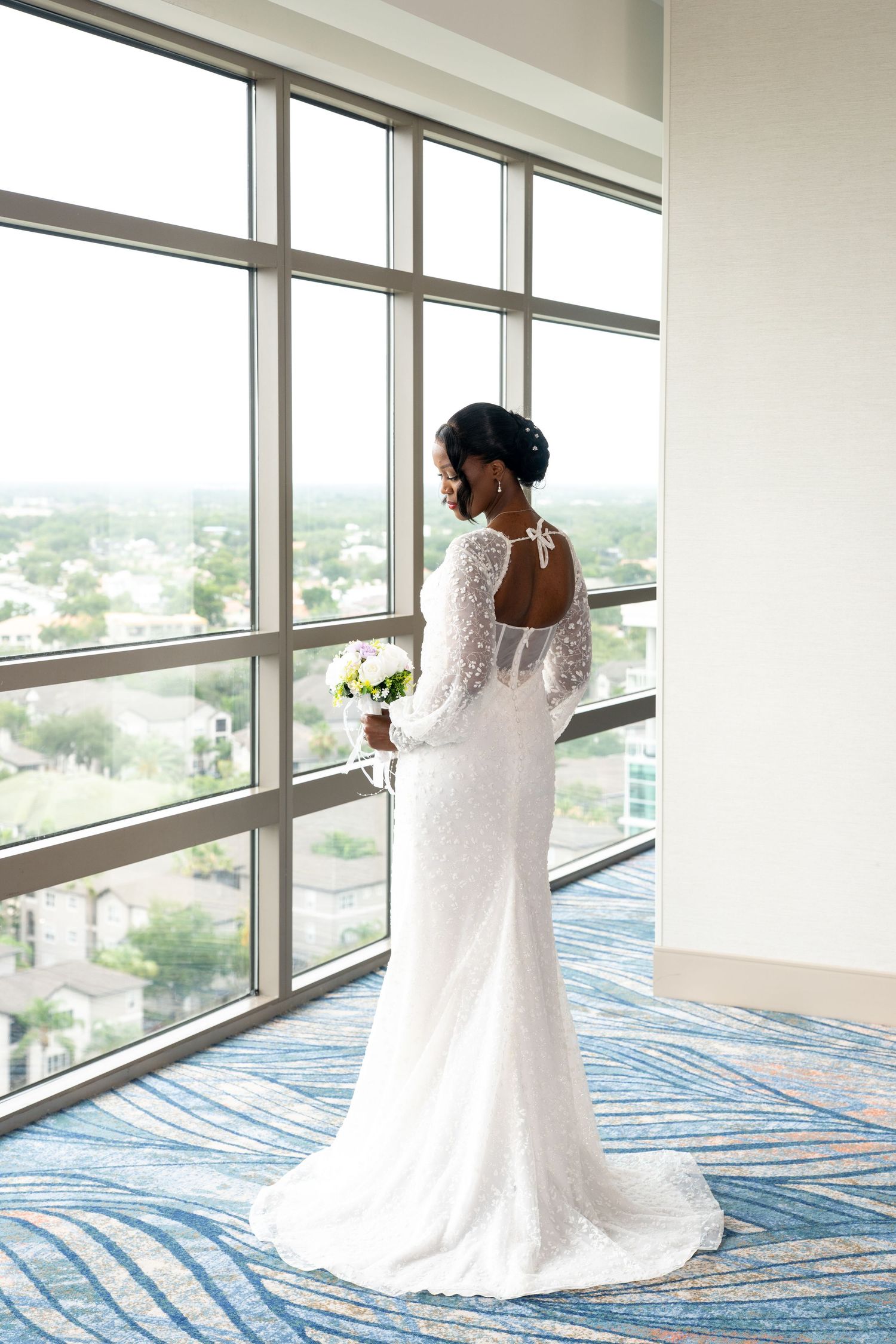 Bride in white lace long-sleeve gown holding bouquet, standing before large windows with city view.