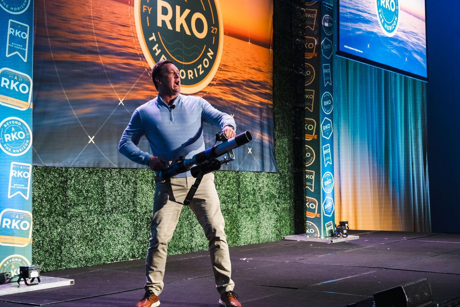 Man on stage at RKO event holding a rifle prop in front of branded backdrop with orange horizon logo.