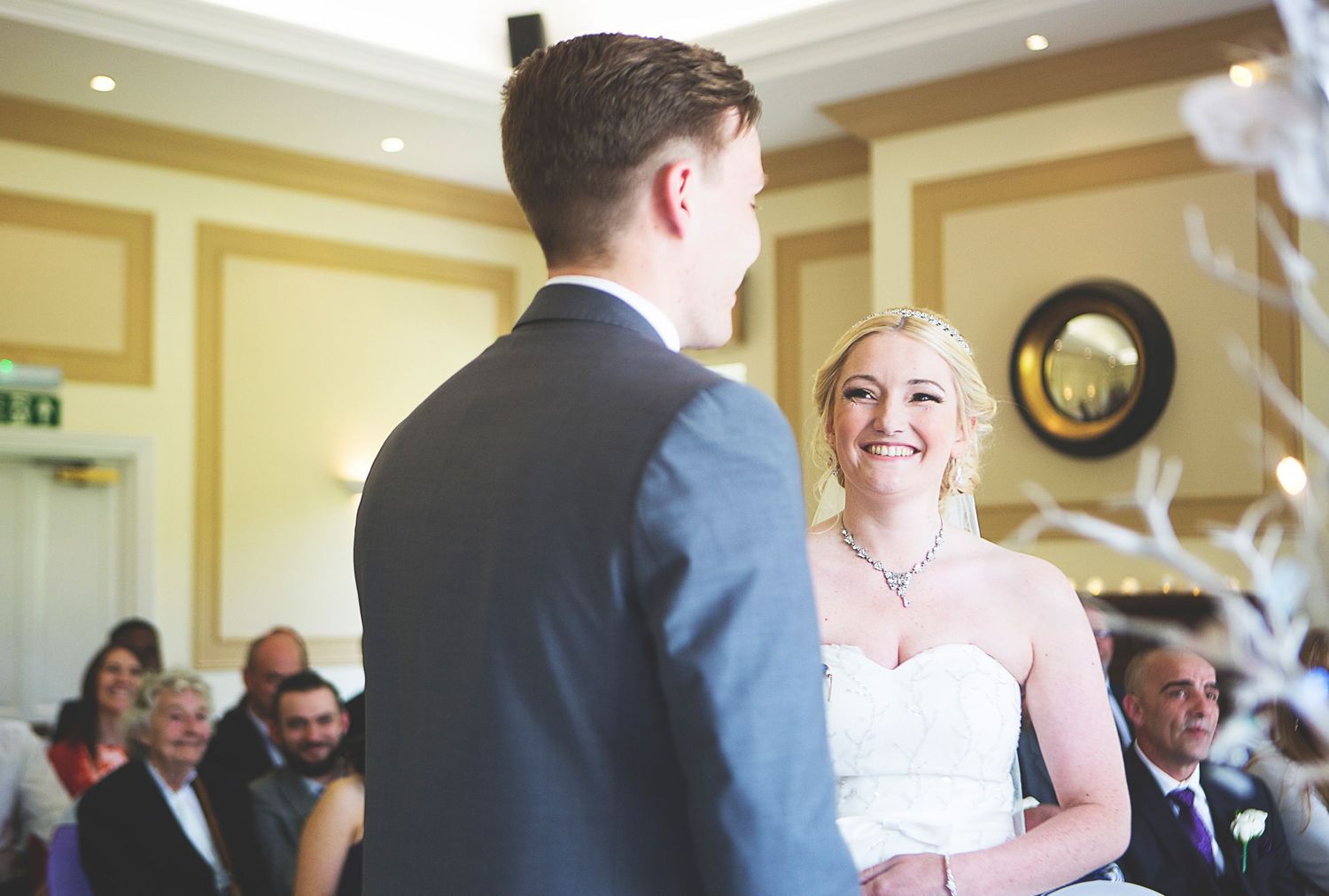 Bride smiling joyfully at groom during indoor wedding ceremony, guests seated in background.