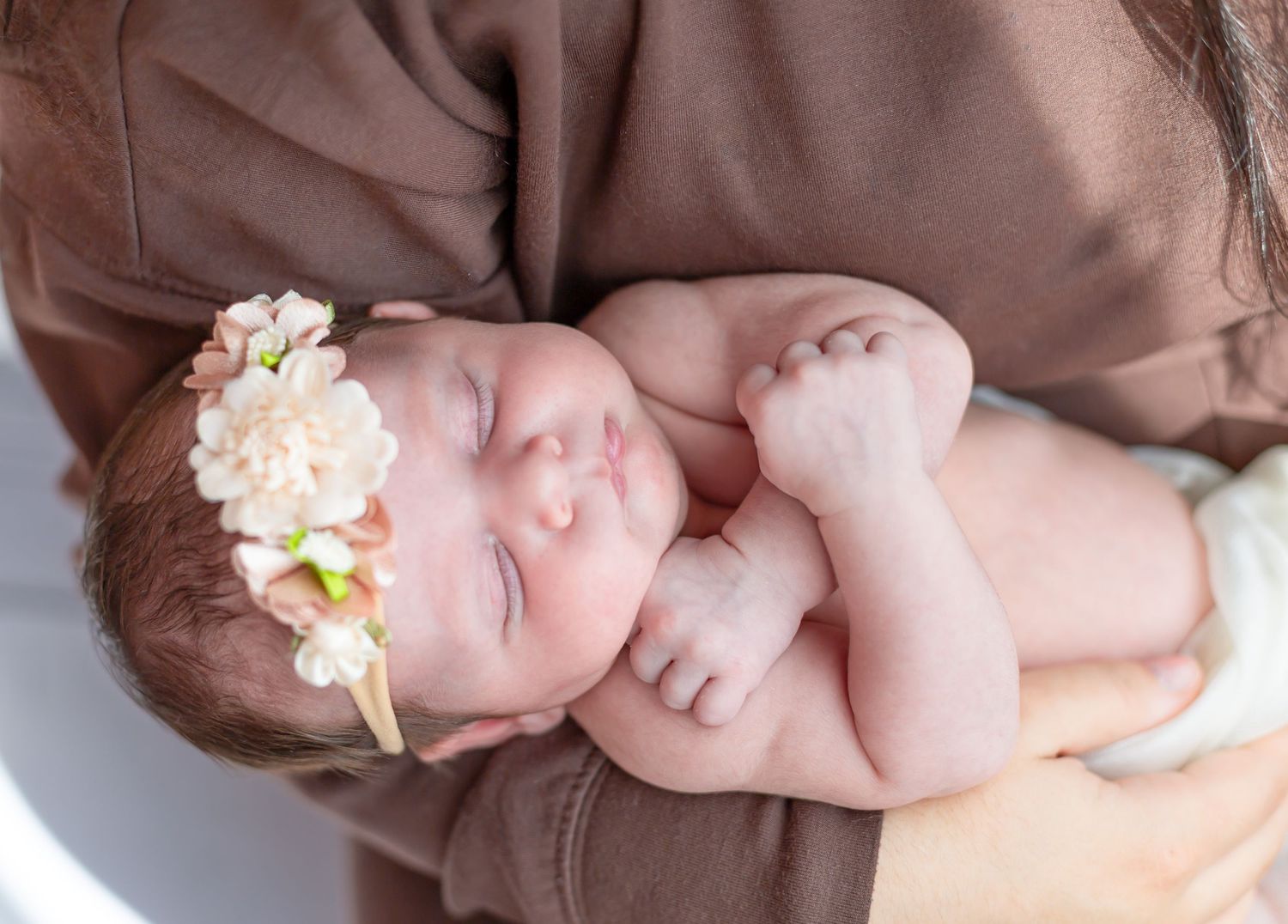 Sleeping newborn baby girl wearing a floral headband, cradled in mother's arms wearing a brown outfit.