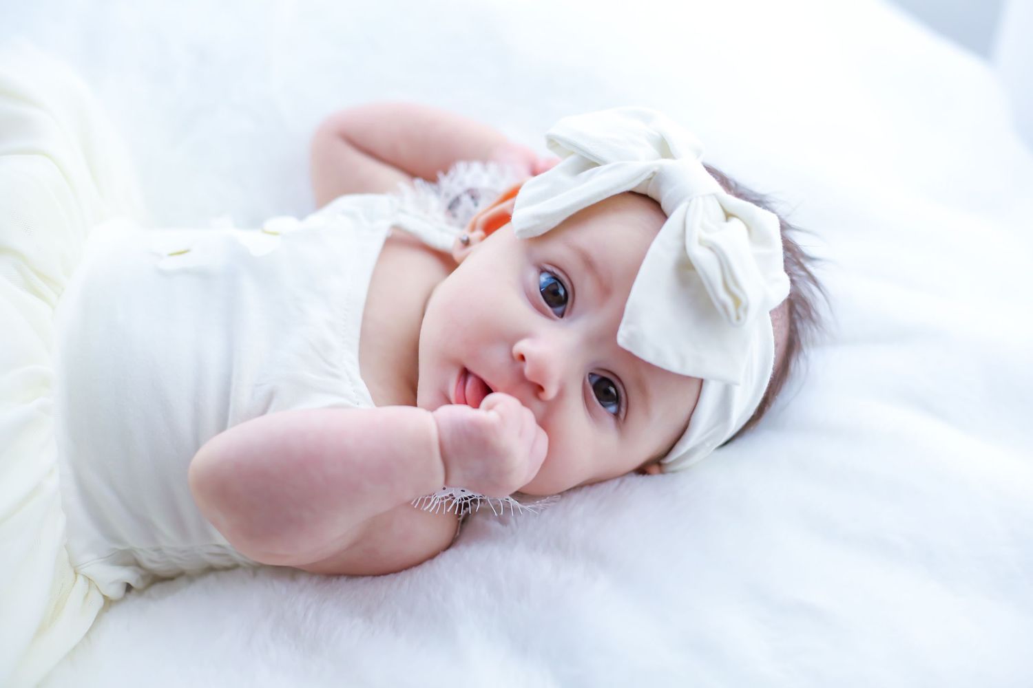 Adorable baby girl in white outfit and bow headband lying on fluffy white blanket, smiling with bright blue eyes.