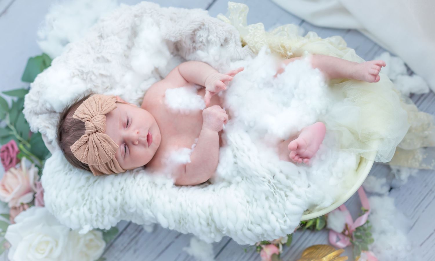 Newborn baby sleeping peacefully in a white fluffy basket with a bow headband, surrounded by soft flowers.