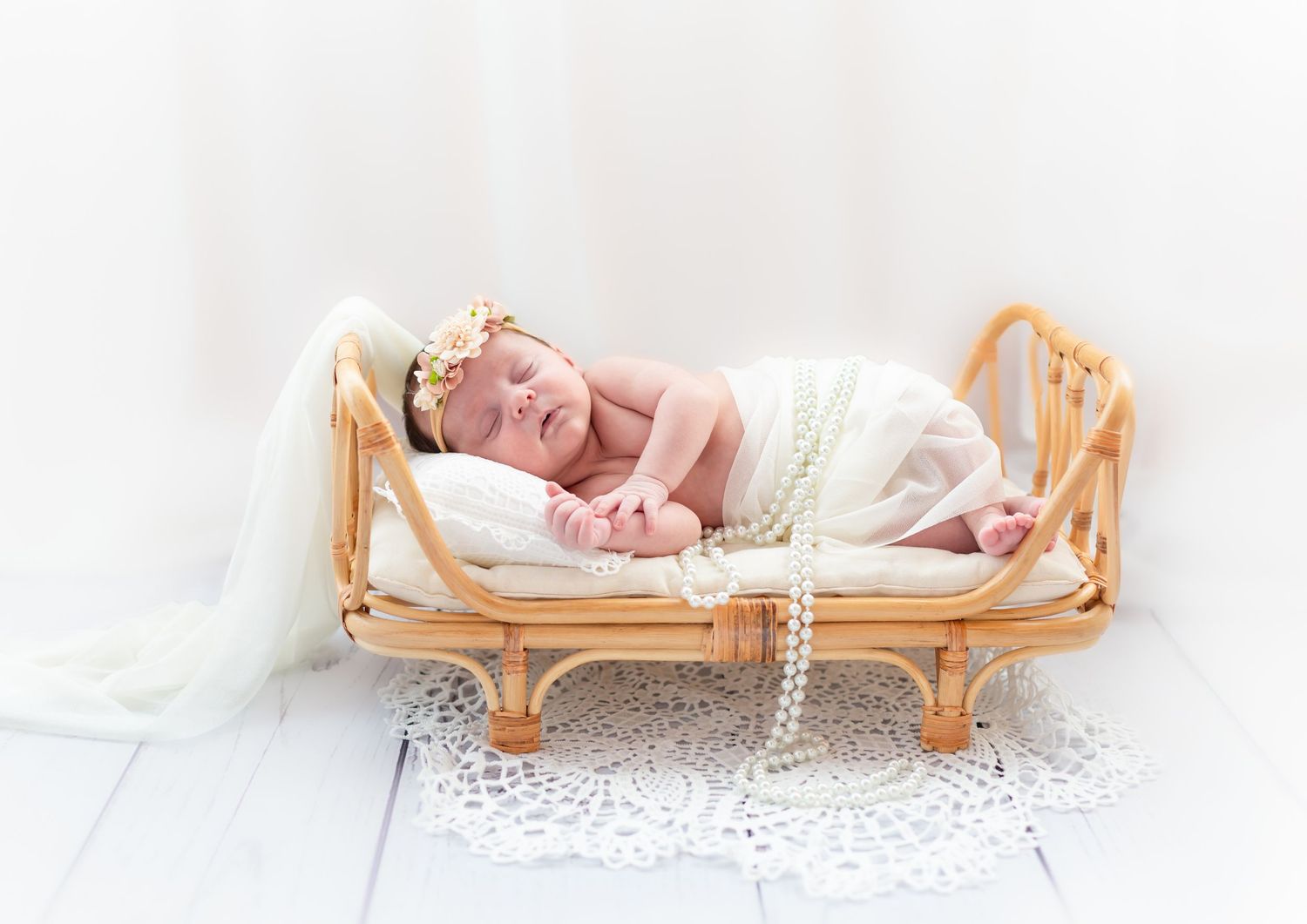 Newborn baby sleeping peacefully on a tiny rattan bed with white lace and pearl decorations.