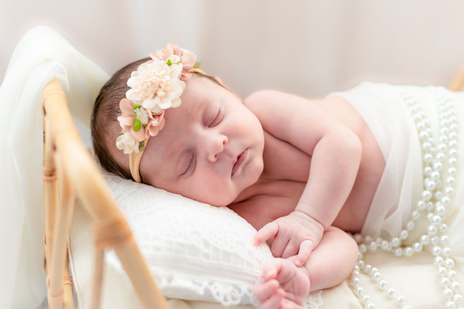 Sleeping newborn baby girl wearing a pink floral headband, nestled in a white basket with pearl necklace decor.