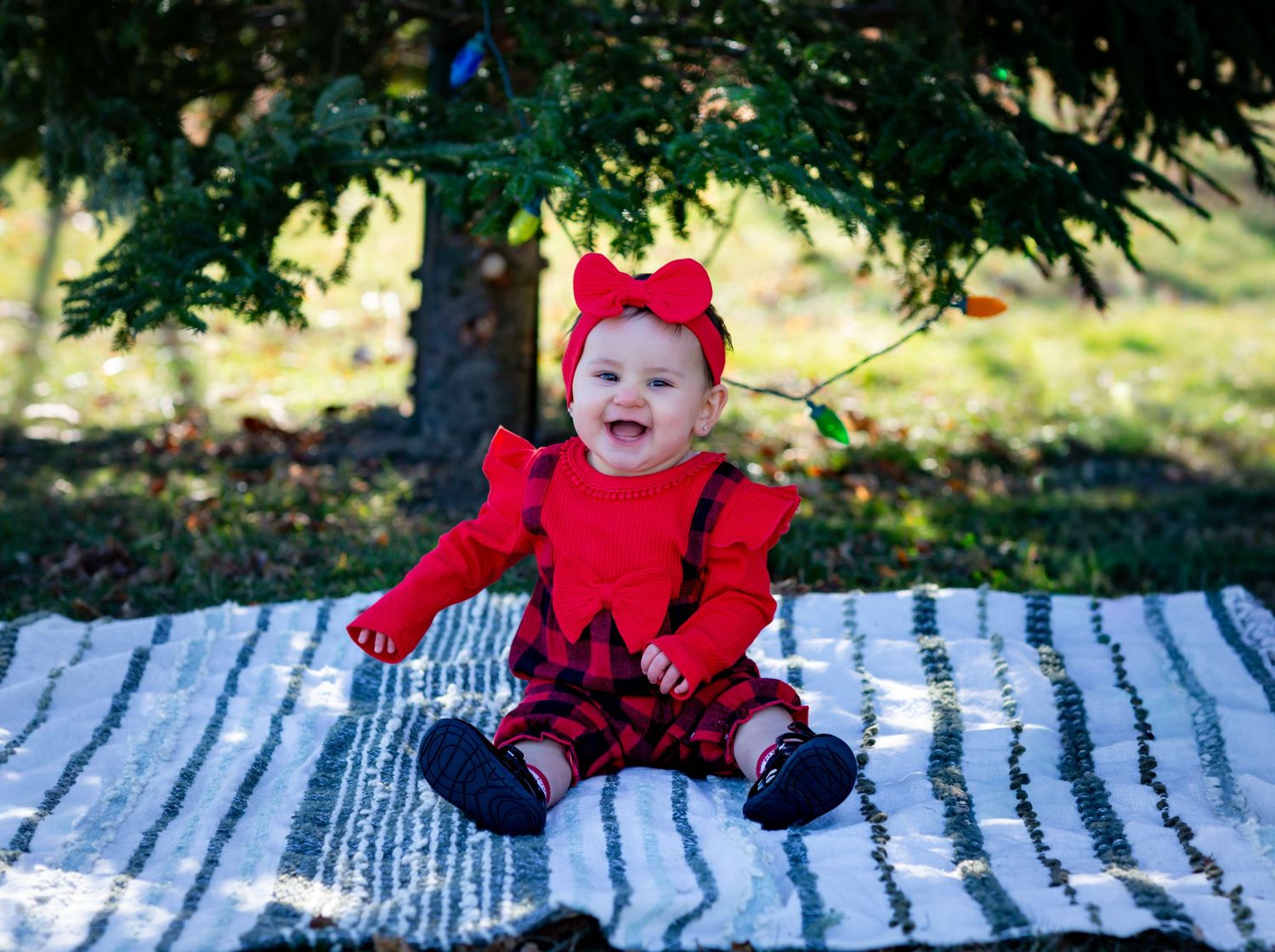 Happy baby girl in red Christmas outfit sitting on blanket under evergreen tree outdoors.