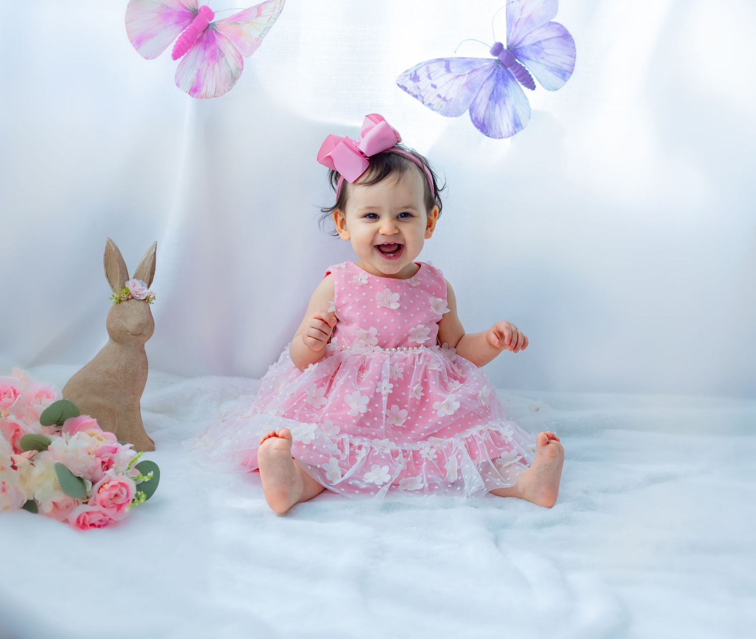 Smiling baby girl in pink dress sits on white blanket with Easter bunny, roses, and butterfly decorations.