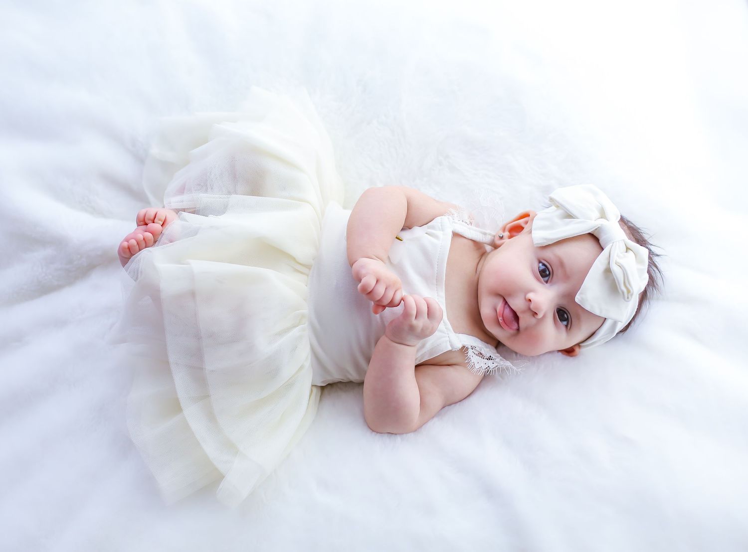Smiling baby girl in white tutu dress and bow headband lying on fluffy white blanket.