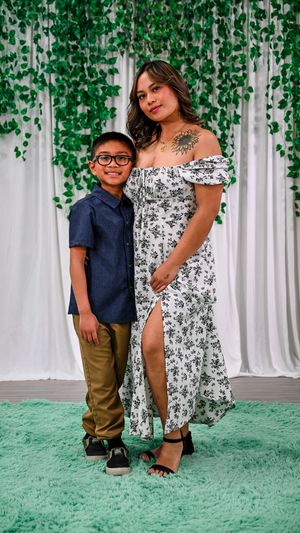 A young boy in glasses and a woman in a floral off-shoulder dress pose together in front of a green vine backdrop.