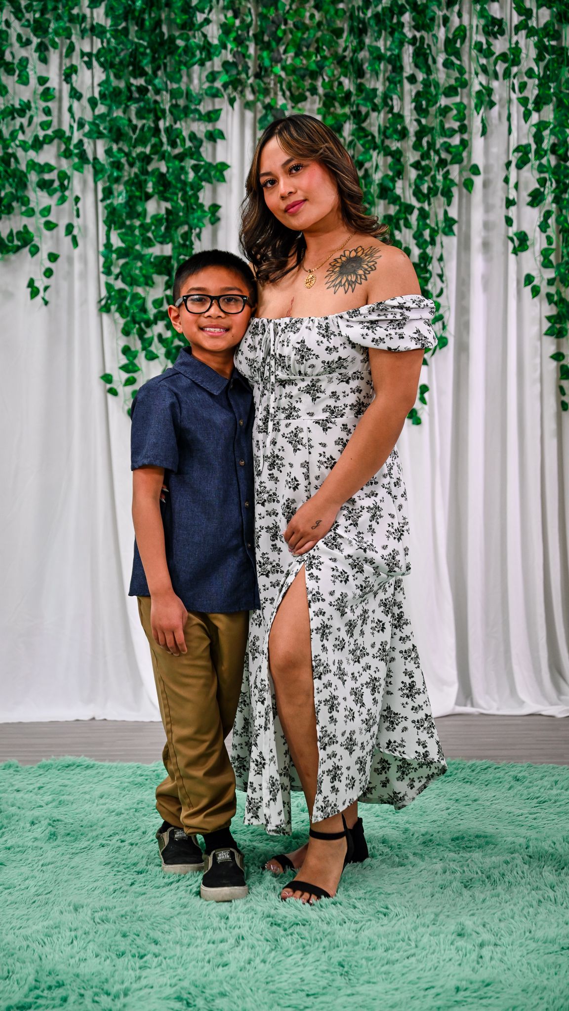 A young boy in glasses and a woman in a floral off-shoulder dress pose together in front of a green vine backdrop.