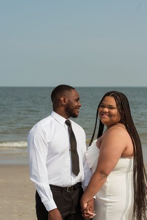 A couple holds hands on a beach, dressed in white wedding attire, smiling at each other with the ocean in the background.