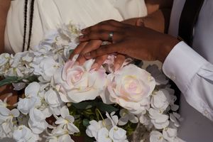 Newlywed couple holding hands over white roses and orchids bouquet, showing wedding rings at ceremony.