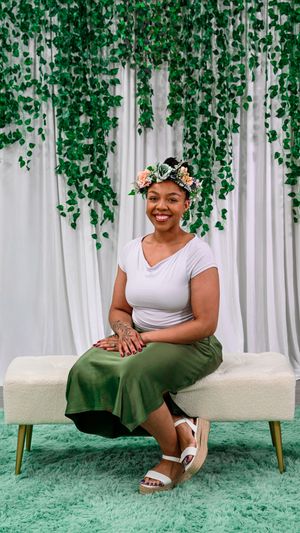 A smiling woman wearing a floral crown and green skirt sits on a white bench with hanging vines backdrop.