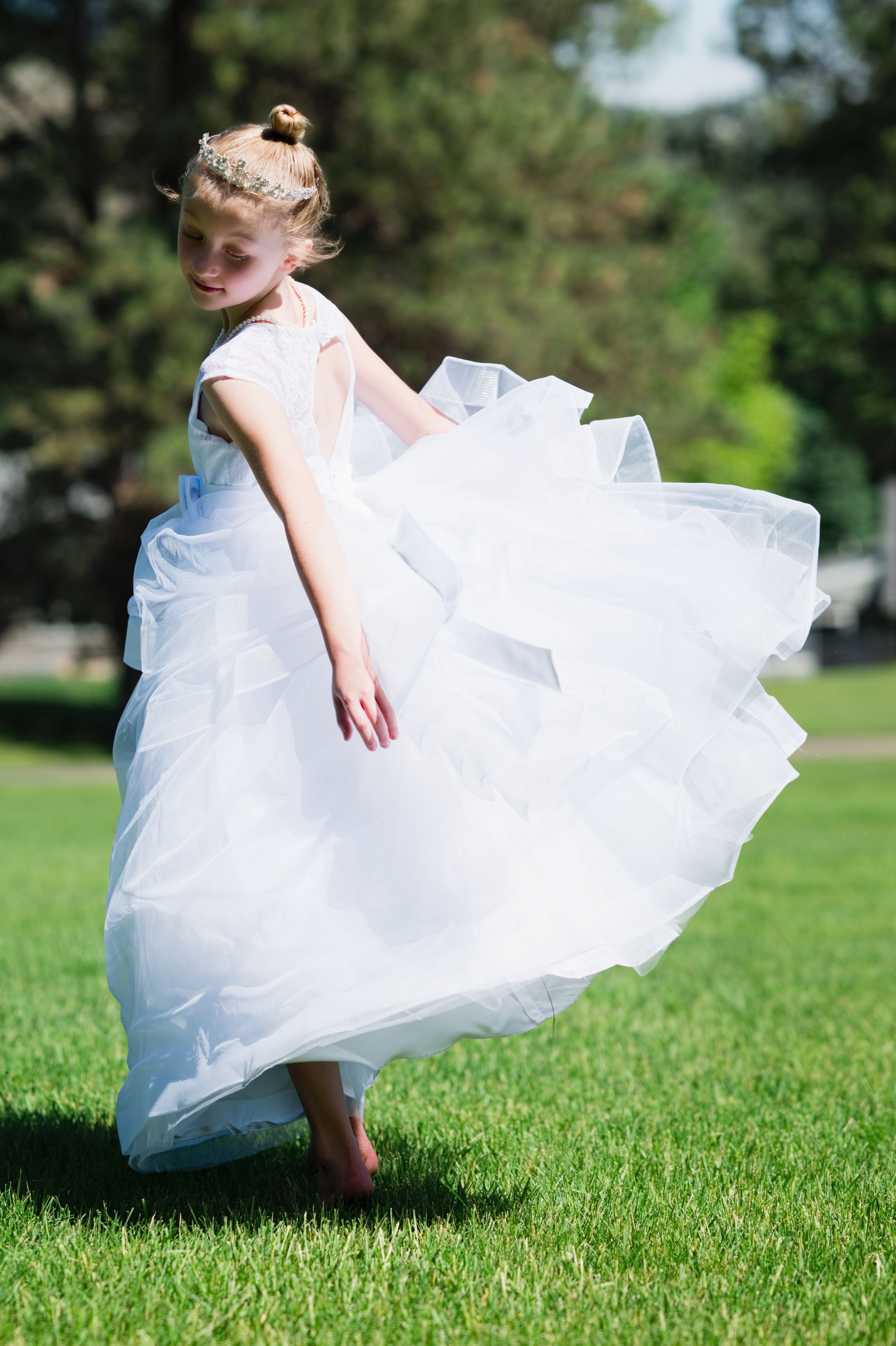 Young girl twirling in white flower girl dress on green grass outdoors.