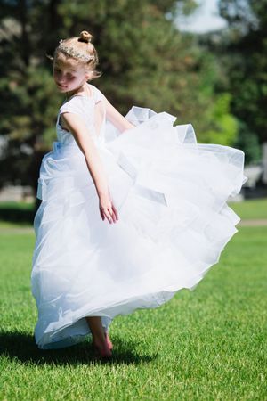 Young girl twirling in white flower girl dress on green grass outdoors.