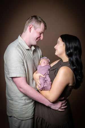 Happy couple smiling at each other while holding their sleeping newborn baby wrapped in purple in a studio portrait.