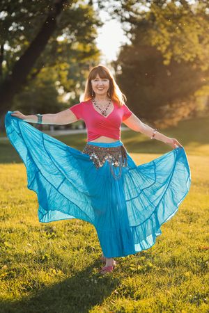 Woman in red top and turquoise belly dance skirt twirling outdoors in sunlit park.