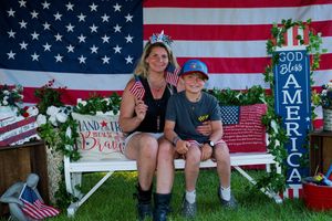 Woman and boy sit on white bench with American flags and God Bless America sign for patriotic photo.
