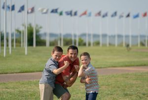 A man playfully wrestles with two young boys on a green lawn with colorful international flags in the background.