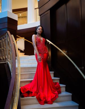 Woman in red mermaid gown with gold beading poses elegantly on marble staircase in luxurious interior.