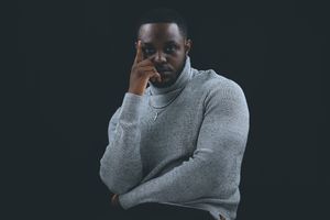 Thoughtful Black man in gray turtleneck sweater posing with hand on face against dark background.