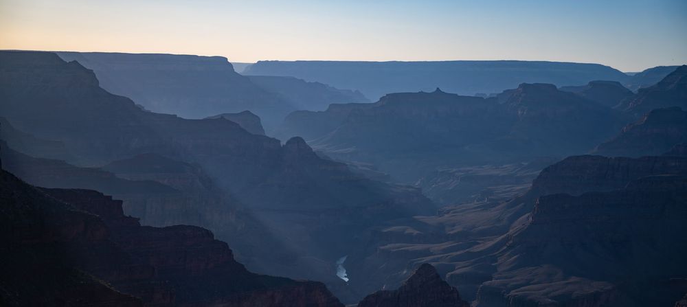 Panoramic twilight view of the Grand Canyon with layered silhouettes and the Colorado River visible below.