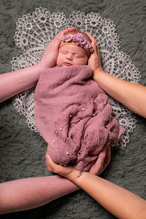 Newborn baby girl swaddled in pink wrap with floral headband, cradled by parents hands on lace doily.