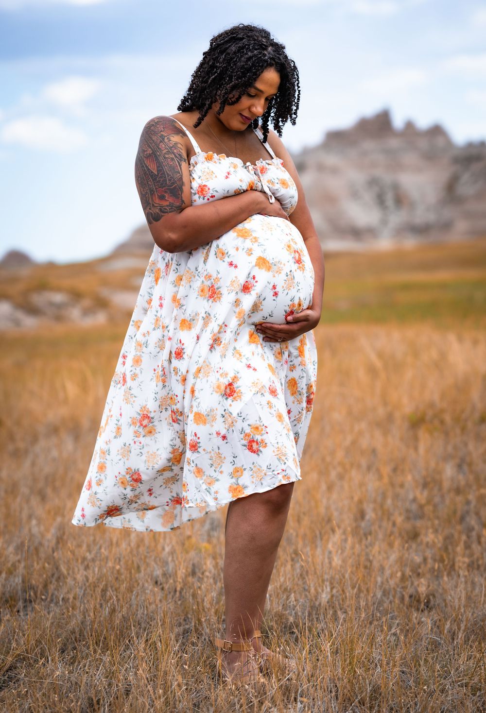 Pregnant woman in floral sundress cradles baby bump in golden field with rocky badlands landscape behind her.