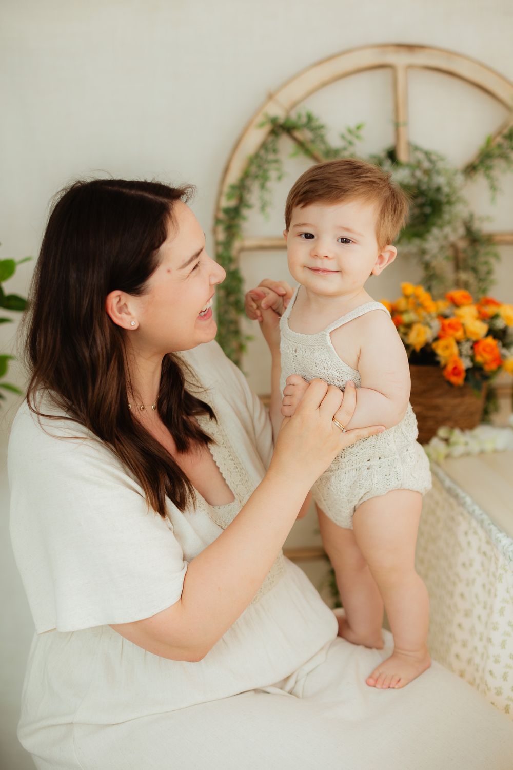 A mother holds her smiling baby in a bright, floral-decorated room, both dressed in light, soft clothing.
