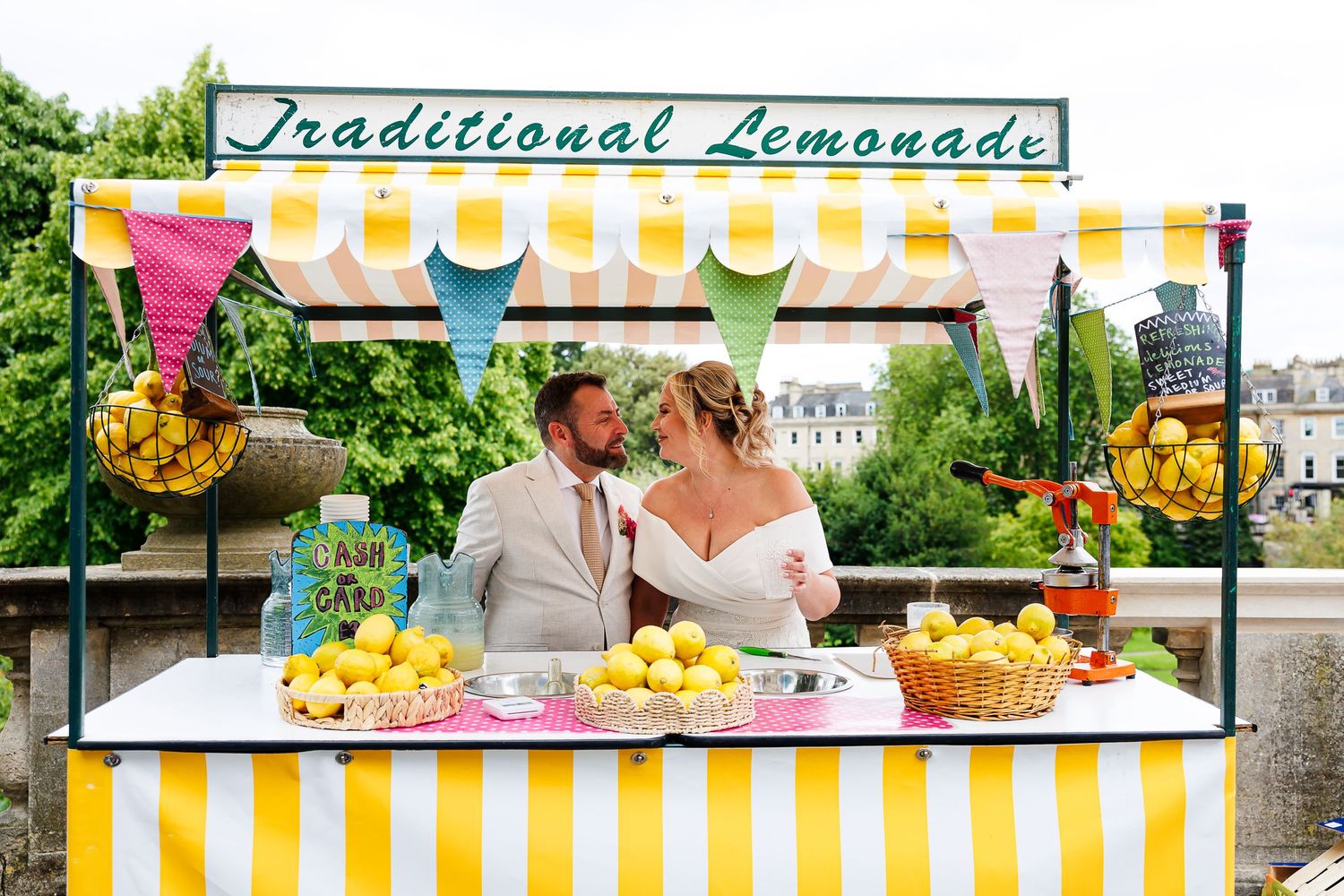 Newlywed couple smiling at a colorful Traditional Lemonade stand with fresh lemons on their wedding day.