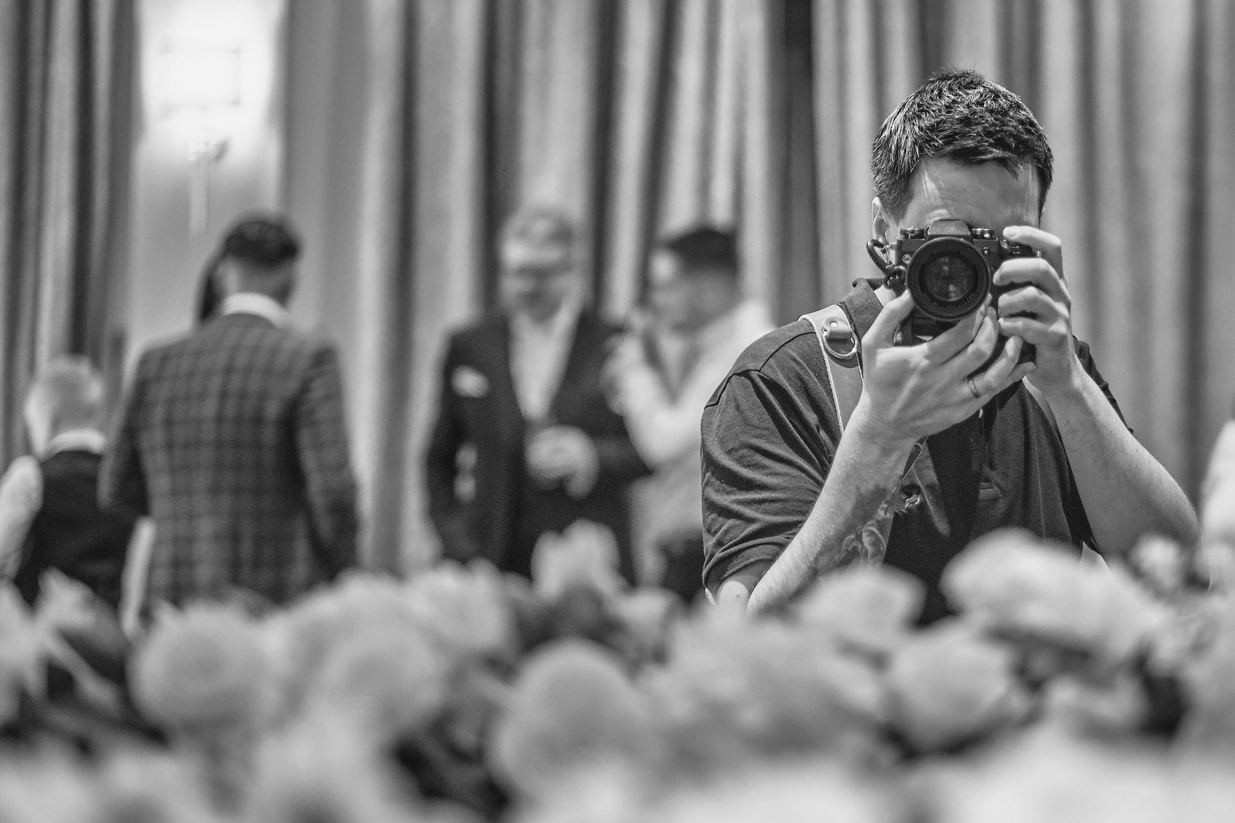 Black and white photo of a photographer capturing images at a formal event with guests in suits in the background.