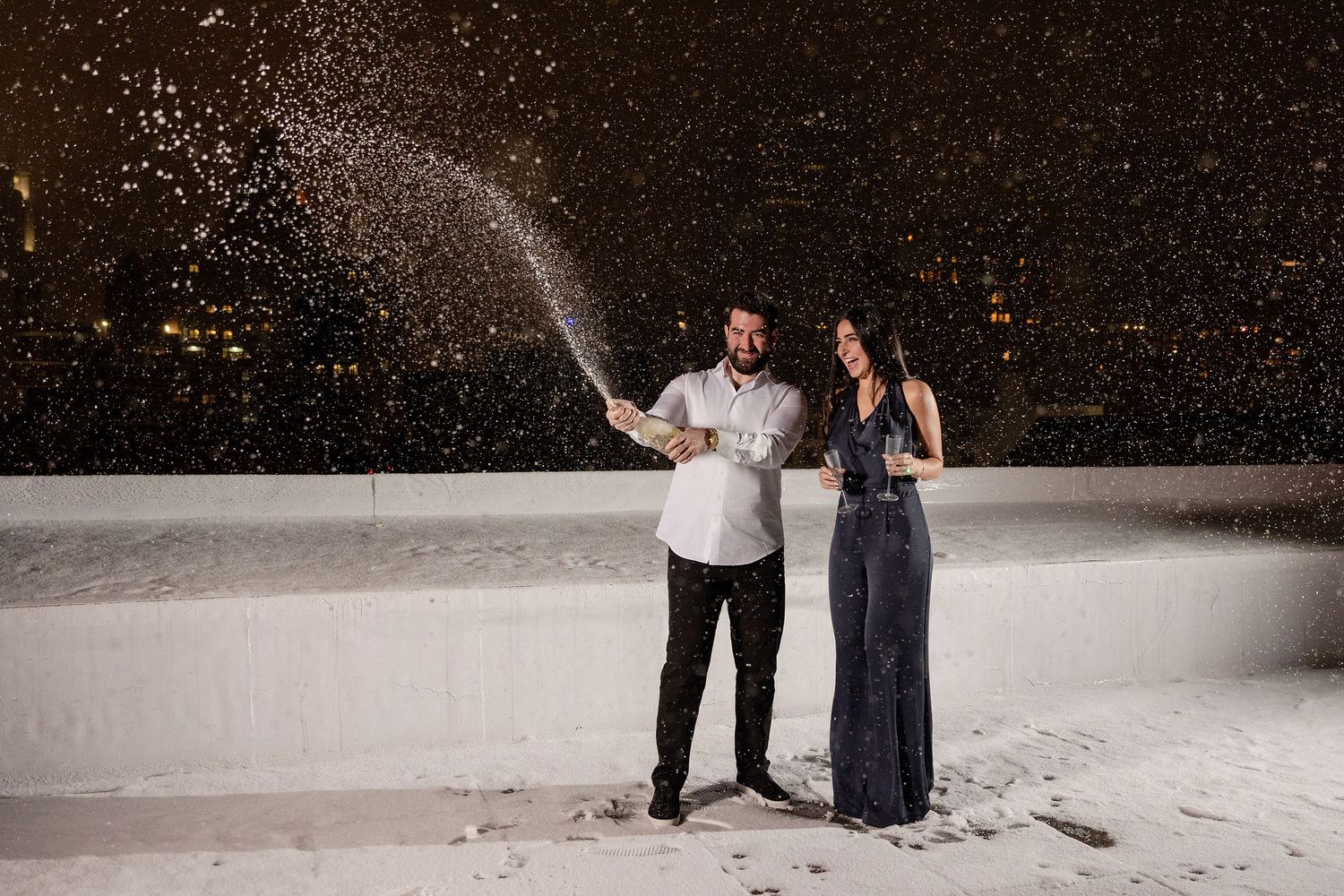 Couple celebrating on snowy rooftop at night, spraying champagne in elegant attire.