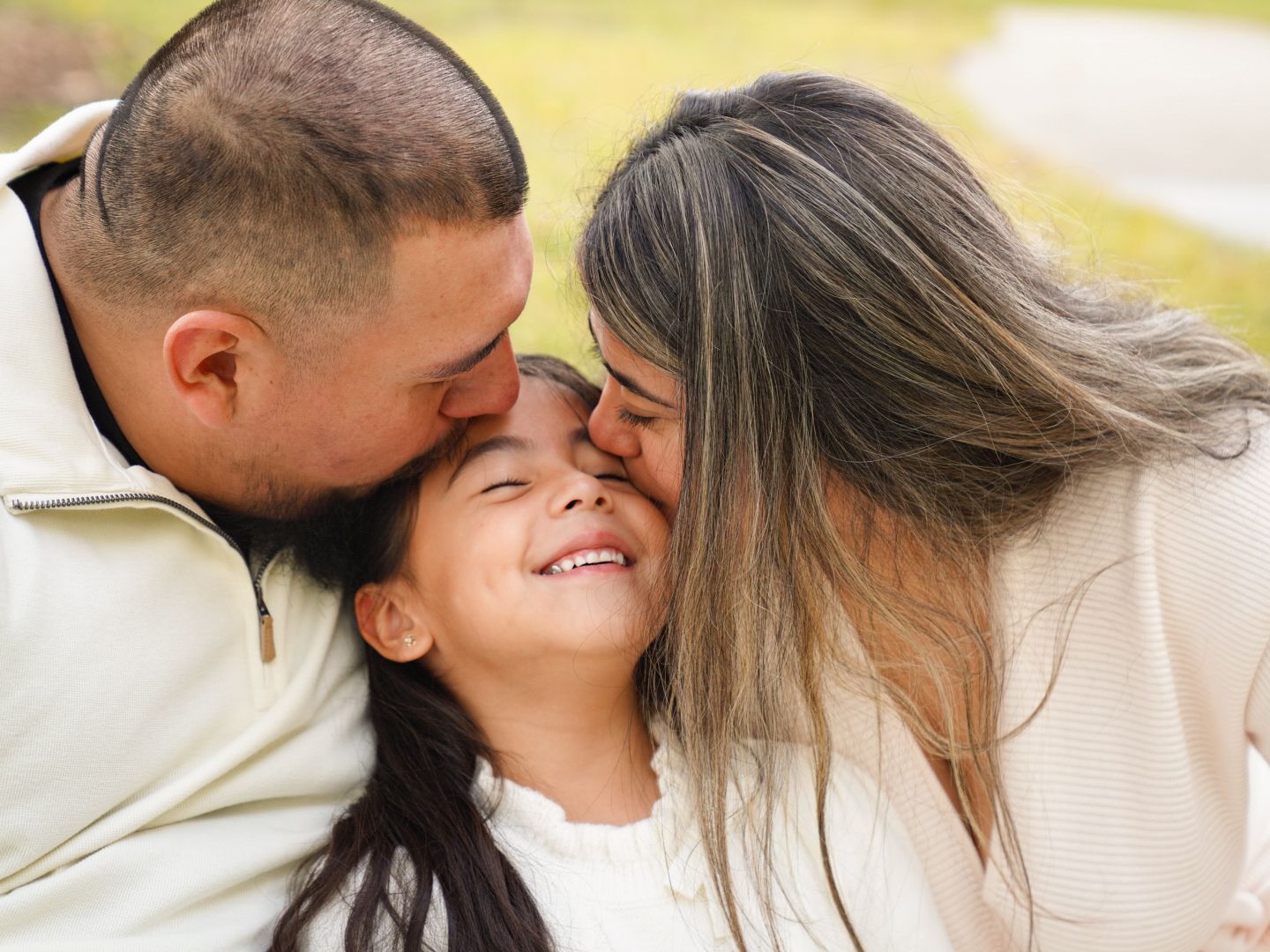 Happy family moment with parents kissing their smiling young daughter on the cheeks outdoors.