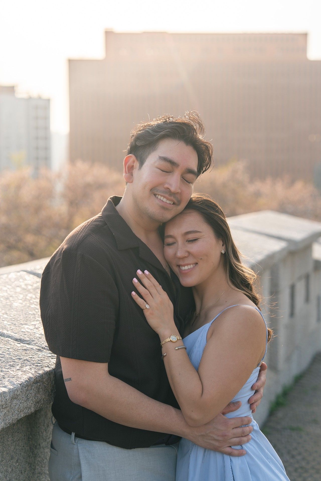 A smiling couple embraces on a rooftop at golden hour, with a city skyline glowing softly in the background.