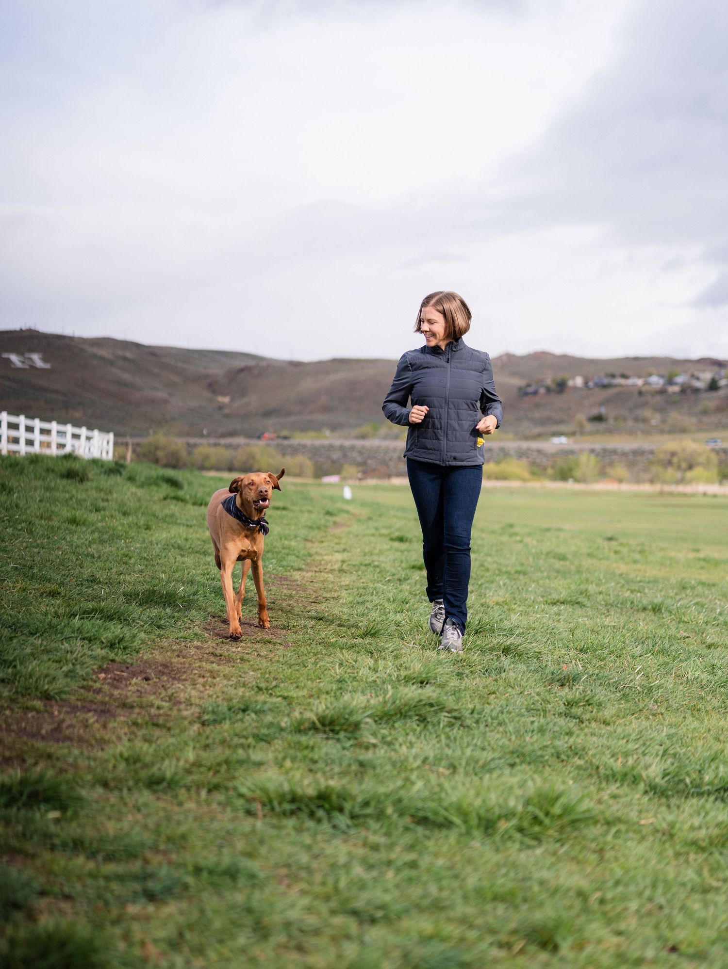 Woman jogging with a brown dog on a green grassy field with rolling hills in the background.