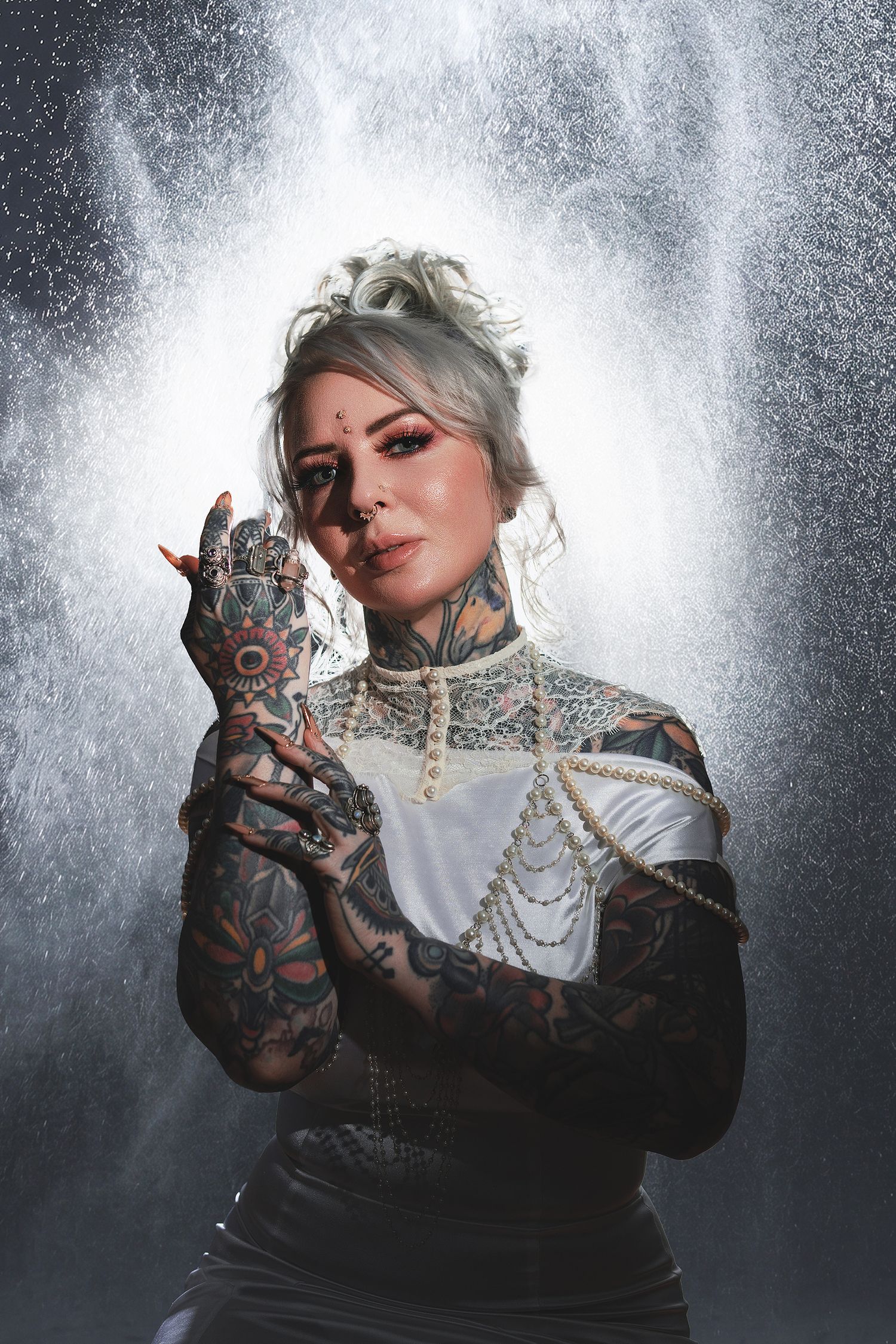 Tattooed woman facing camera with hands framing face, draped pearl chains, backlit water column behind her, creative portrait photography
