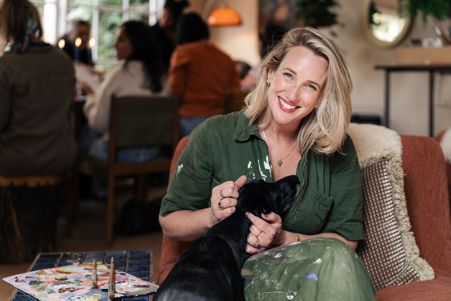 A smiling blonde woman holds a small black puppy while sitting on a couch in a cozy cafe.

Strange Bird Studio