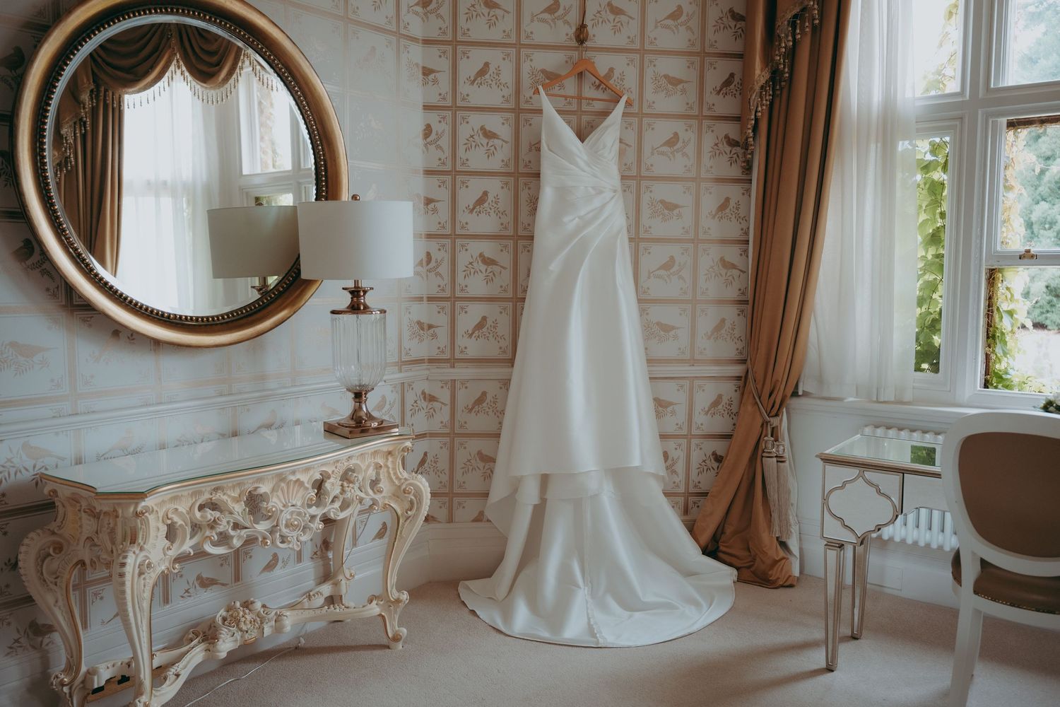 Elegant white wedding dress displayed on a hanger in a luxurious room with ornate mirror and vintage console table.