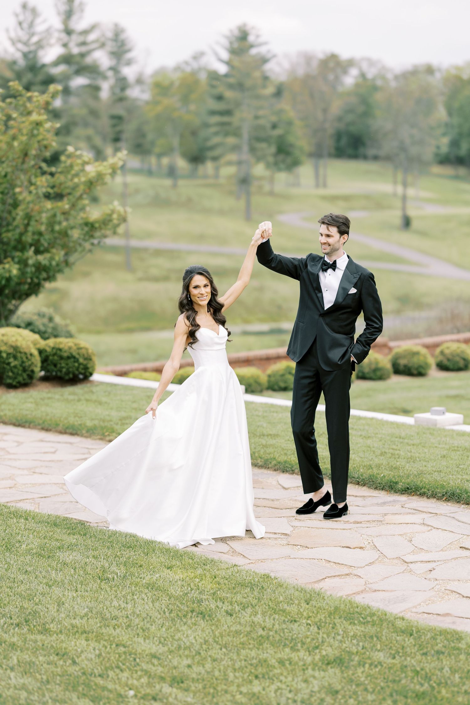 Bride in white gown twirls as groom in black tuxedo raises her hand on a stone path surrounded by lush greenery.