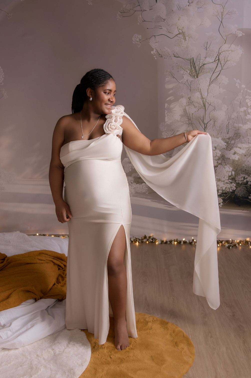 Pregnant woman in elegant white one-shoulder gown with floral detail and flowing sheer fabric, posing in a softly lit studio.
