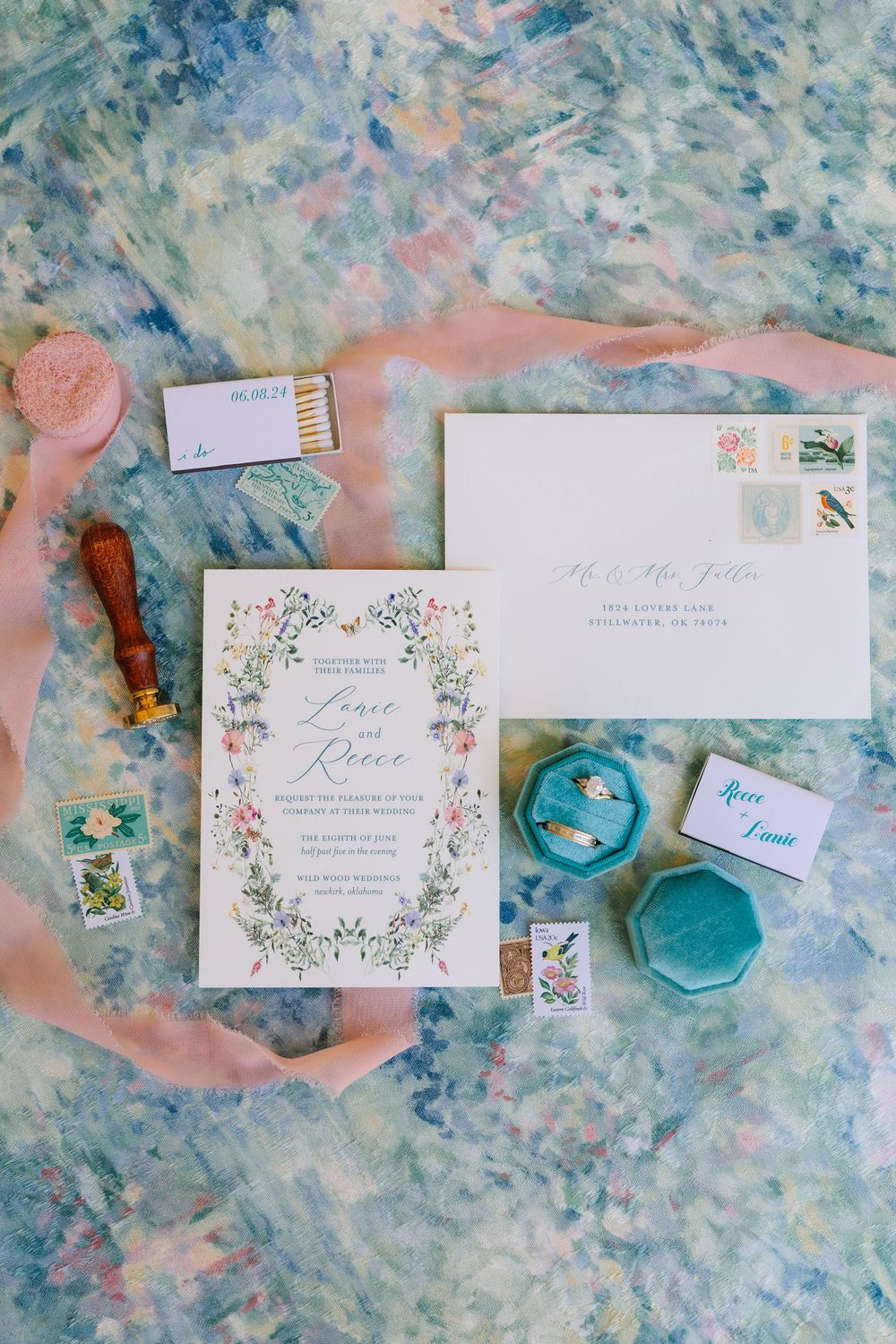 Elegant wedding invitation suite with floral border, wax seal, velvet ring box, vintage stamps, and pink ribbon on watercolor background.