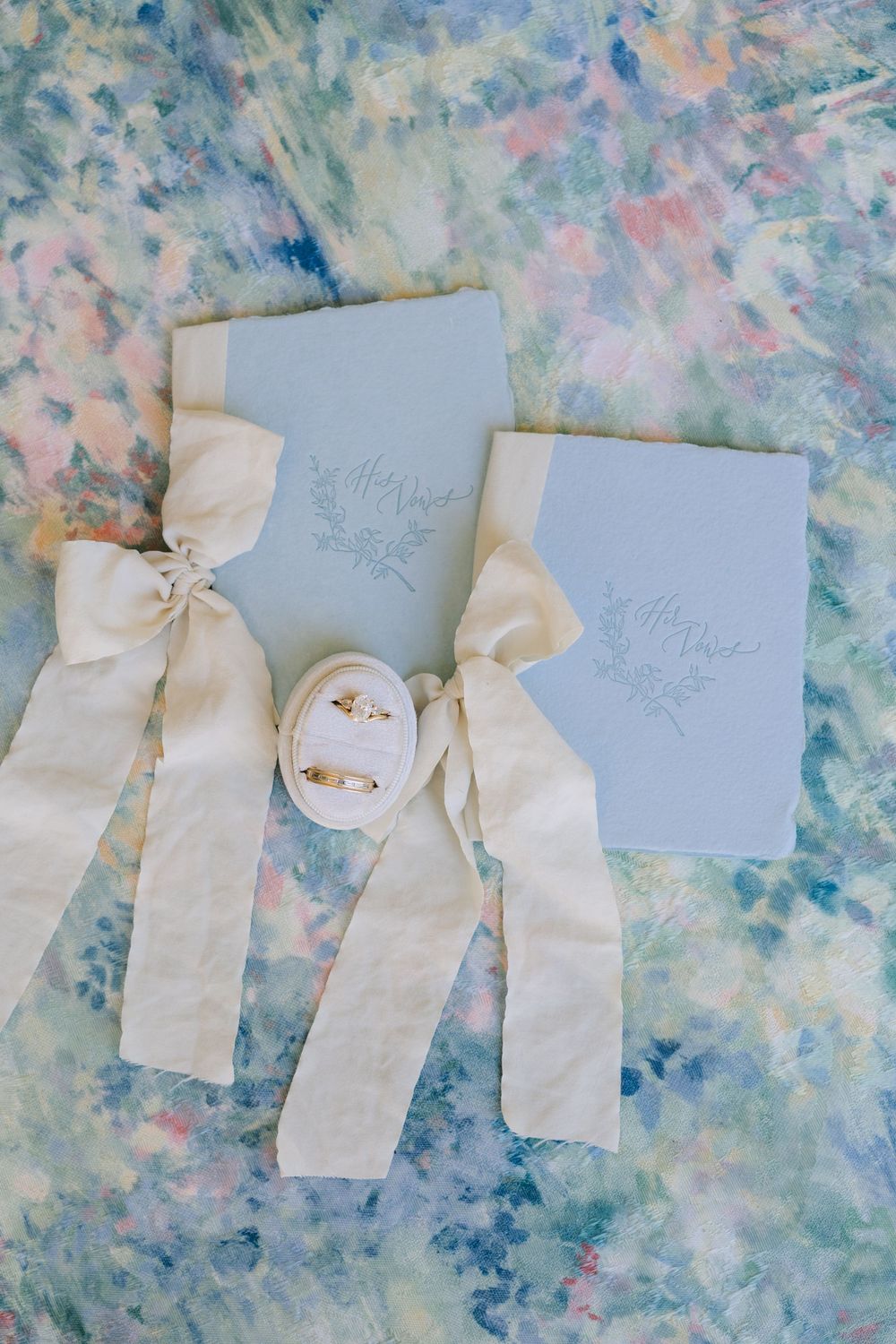 Blue wedding vow books with cream silk ribbon bows and gold rings in velvet box on watercolor background.