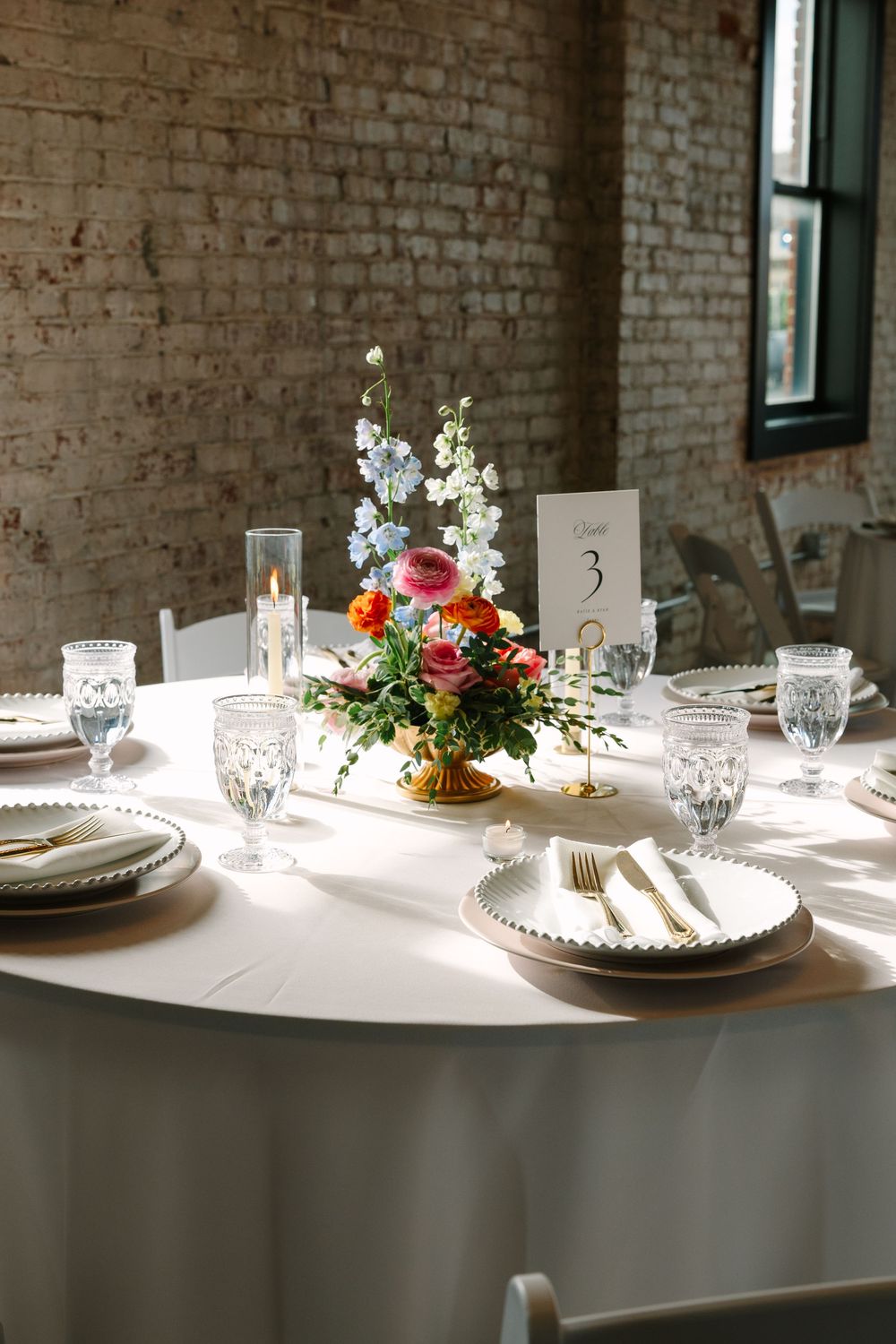 Elegant wedding reception table 3 with floral centerpiece, crystal glasses, gold cutlery, and white linens in a brick venue.