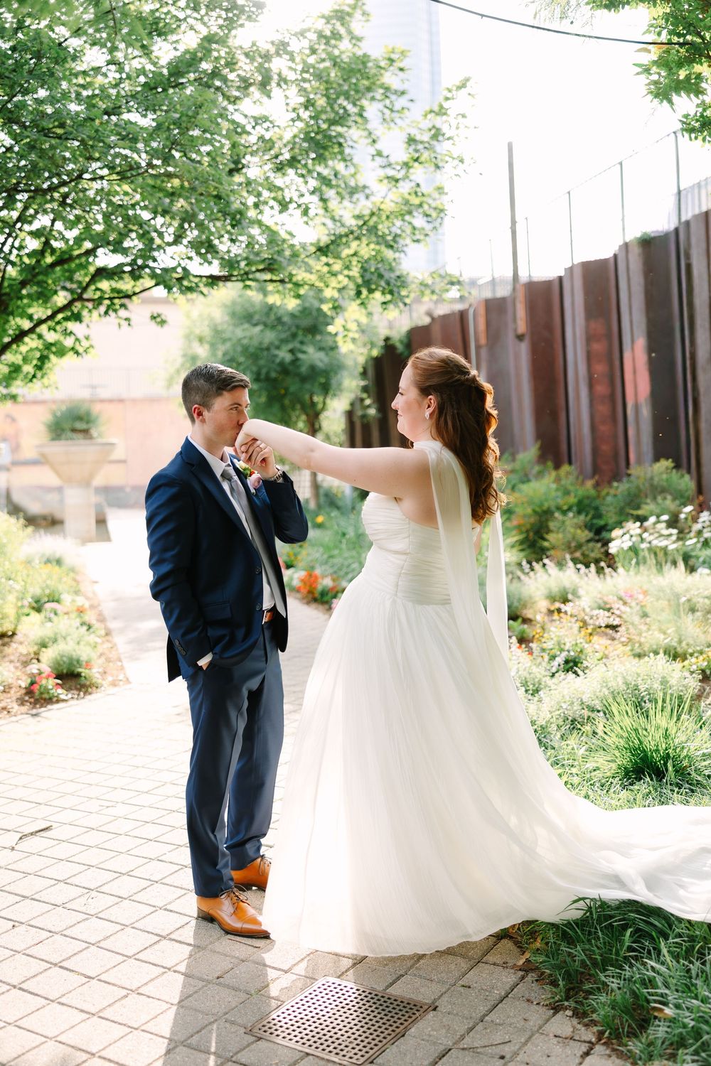 Groom kisses bride's hand in a sunlit garden, bride in white strapless gown, groom in navy suit with tan shoes.