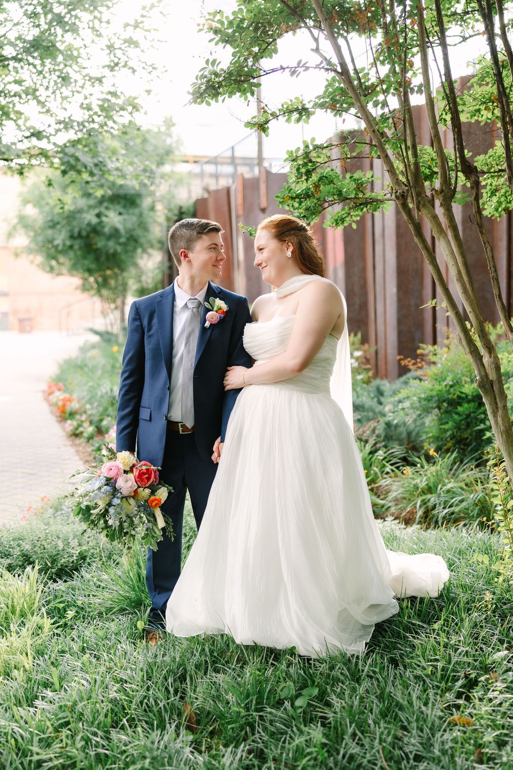 A bride in a white gown and groom in a navy suit share a loving glance outdoors in a lush garden setting.