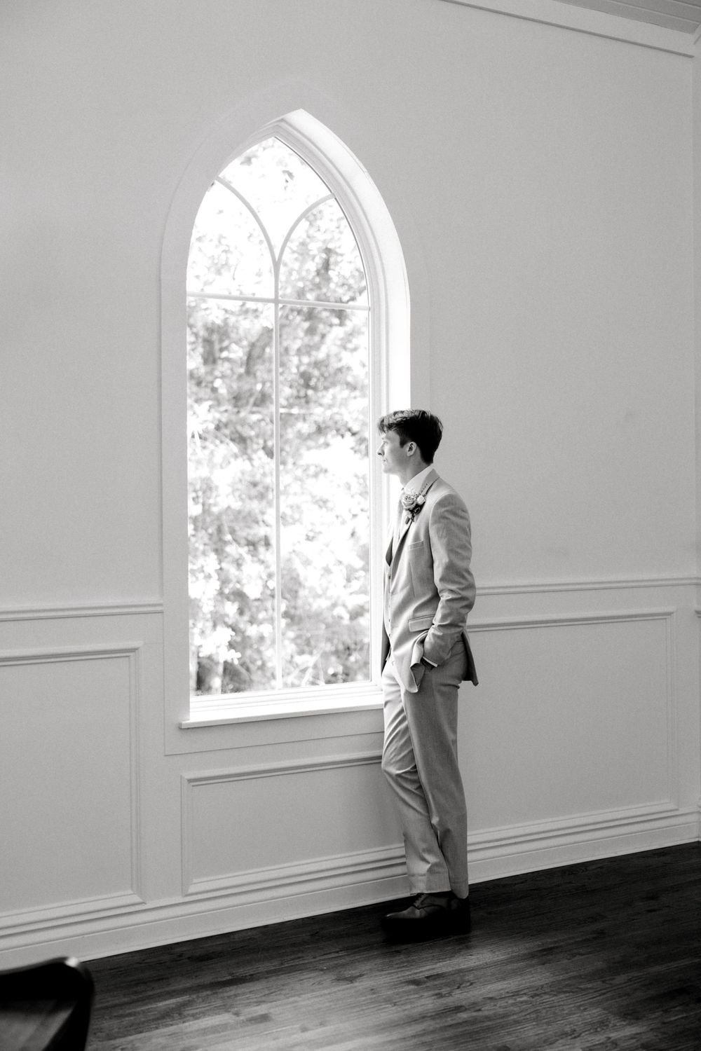 Groom in suit standing by tall arched chapel window in black and white wedding portrait.