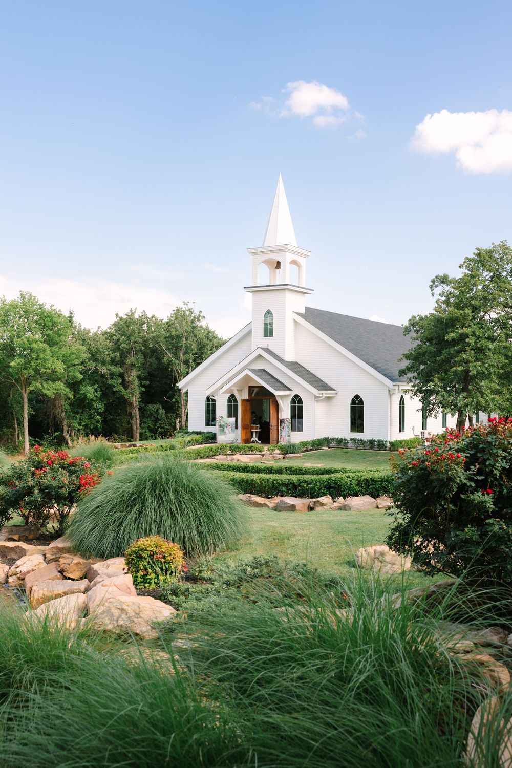 White chapel with steeple surrounded by lush gardens, red roses, and stone landscaping under a blue sky.
